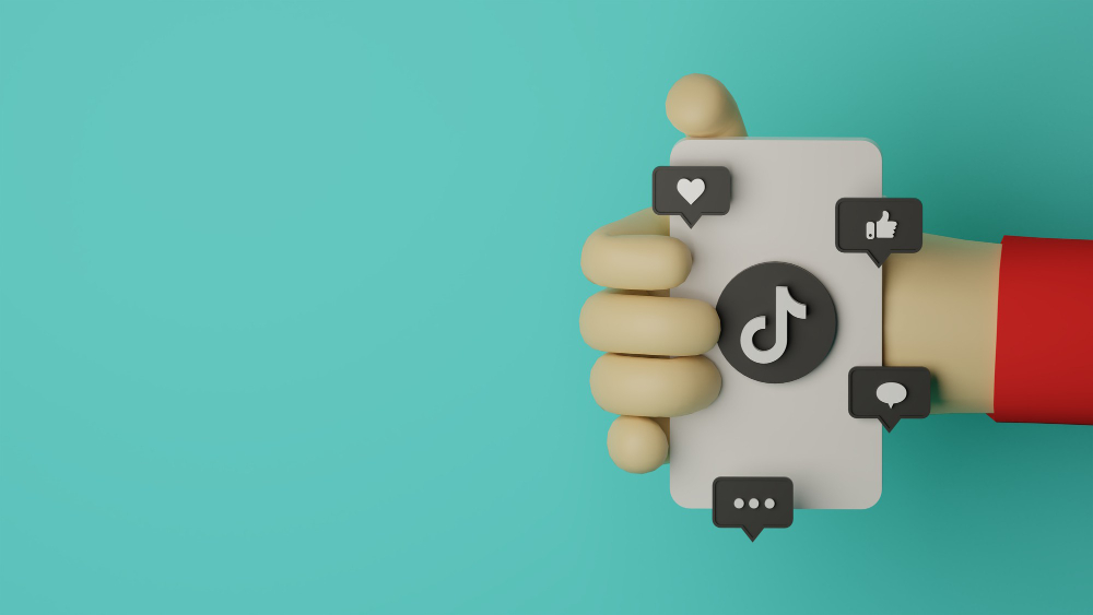 Ręka trzymająca kontur telefonu z widocznym logiem TikTok i interakcji social media (like, follow, komentarze)